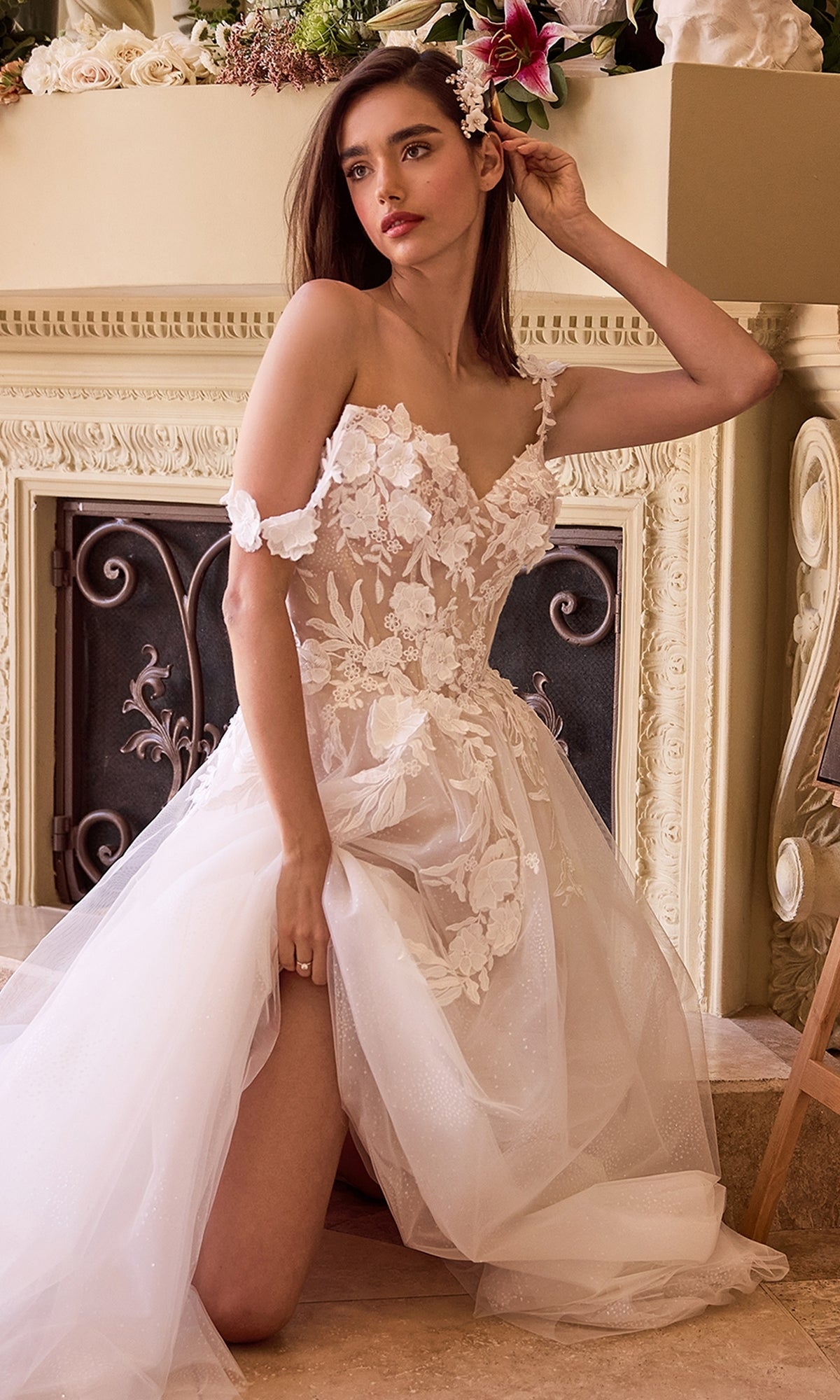 A woman kneels indoors by a fireplace, touching her hair, wearing the Ladivine CDS498W—a long off-white bridal gown with off-the-shoulder floral lace for a romantic, elegant look.